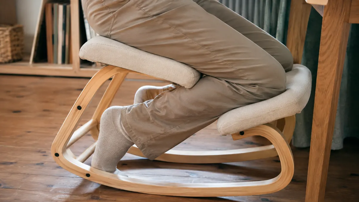 Close-up of a man's knee resting against the padded shin rest of the NYPOT kneeling chair while seated at a desk