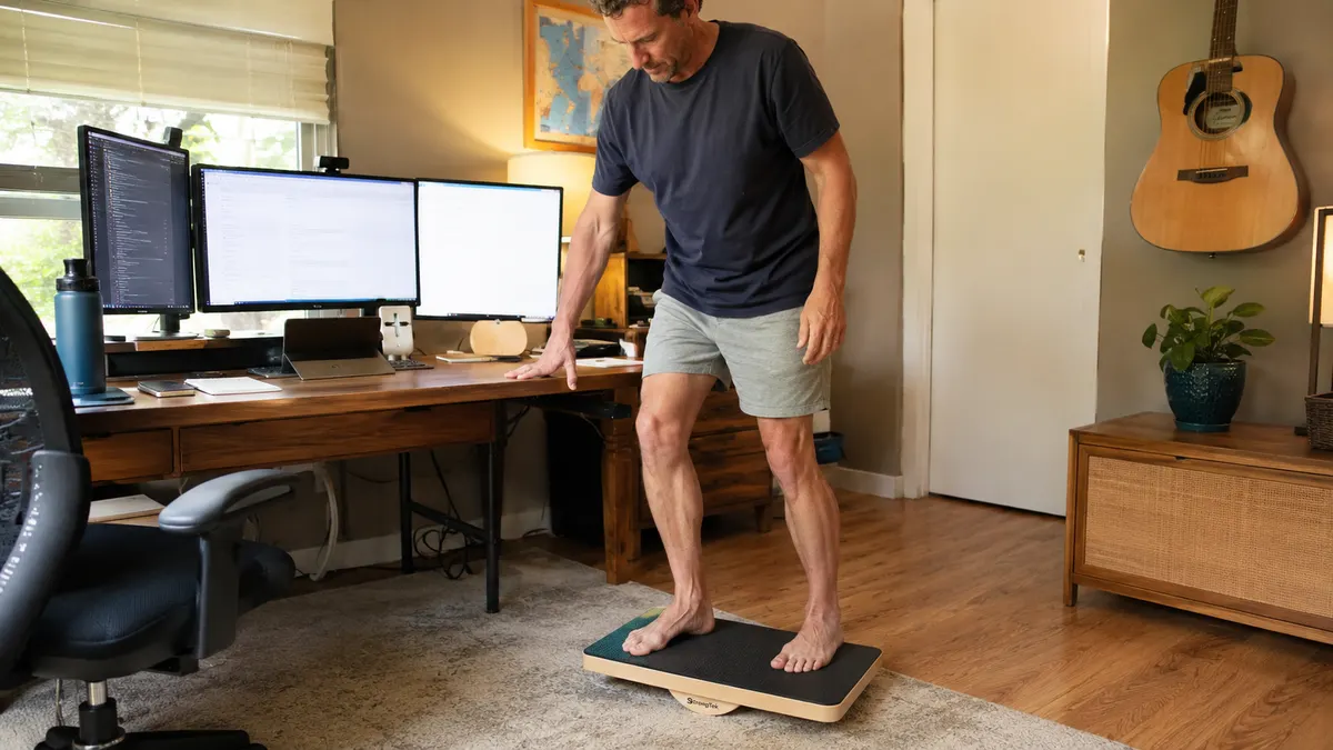Man in socks carefully balancing on a wooden wobble board with one hand on the desk for support